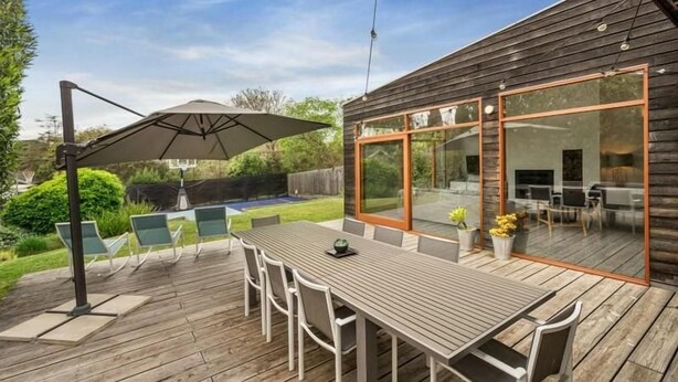 Terraza de madera con mesa y sillas, un área de patio con cerramiento de vidrio y vista a una piscina.