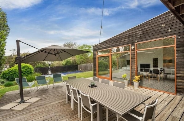 Terraza de madera con mesa y sillas, un área de patio con cerramiento de vidrio y vista a una piscina.