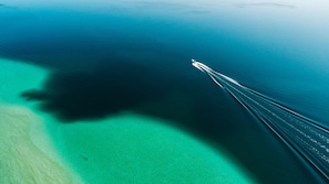 An aerial view of a boat creating a wake in turquoise waters, with a clear demarcation between the sandy beach and the ocean.