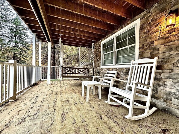 A covered porch with a wooden ceiling, stone walls, and a window with blinds. There are two rocking chairs and a table.
