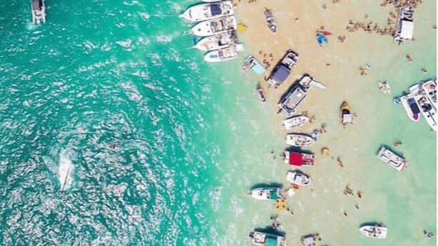 An aerial view of a beach with numerous boats anchored near the shore, a sandy beach, and clear turquoise water. - 3BDR Home by Torch Lake Sandbar 11931 (Rapid City)