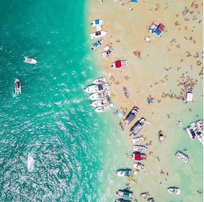 An aerial view of a beach with numerous boats anchored near the shore, a sandy beach, and clear turquoise water.