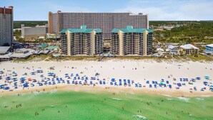 Um hotel à beira-mar com vários edifícios altos, uma praia arenosa com muitos guarda-sóis e uma vista clara do oceano.