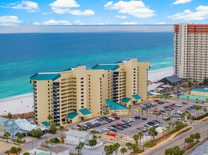 A multi-story hotel with a parking lot, beachfront view, and a large building in the background.