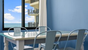 Uma área de refeições com mesa de vidro e cadeiras de metal, uma janela grande com vista para o oceano e um edifício com varandas.