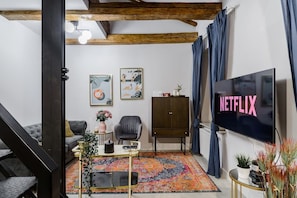 Una sala de estar con un televisor mostrando 'NETFLIX', un sofá, una silla, un mueble aparador y una alfombra colorida.