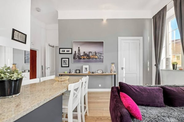 A modern living room with a grey sofa, a kitchen area with a dining table, and a cityscape artwork on the wall.
