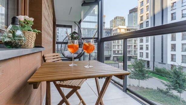 Un balcon avec une table en bois, deux verres de boisson à l’orange, un panier en osier rempli de fleurs et un fauteuil suspendu.