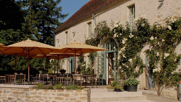 An outdoor seating area with tables and chairs under umbrellas, a stone wall, and a building with a stone facade and green shutters. - Les Deux Ponts (Pierre-Perthuis)
