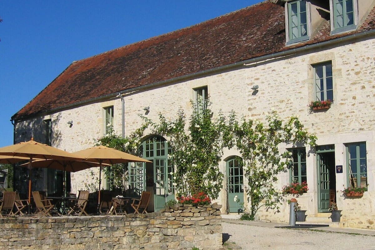 Ein Gebäude aus Stein mit einem Ziegeldach, Außenplätzen mit Sonnenschirmen und grünen Fensterläden.