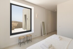 Quarto moderno com uma grande janela, uma cama com roupa de cama branca, dois banquinhos de vime e um espelho.