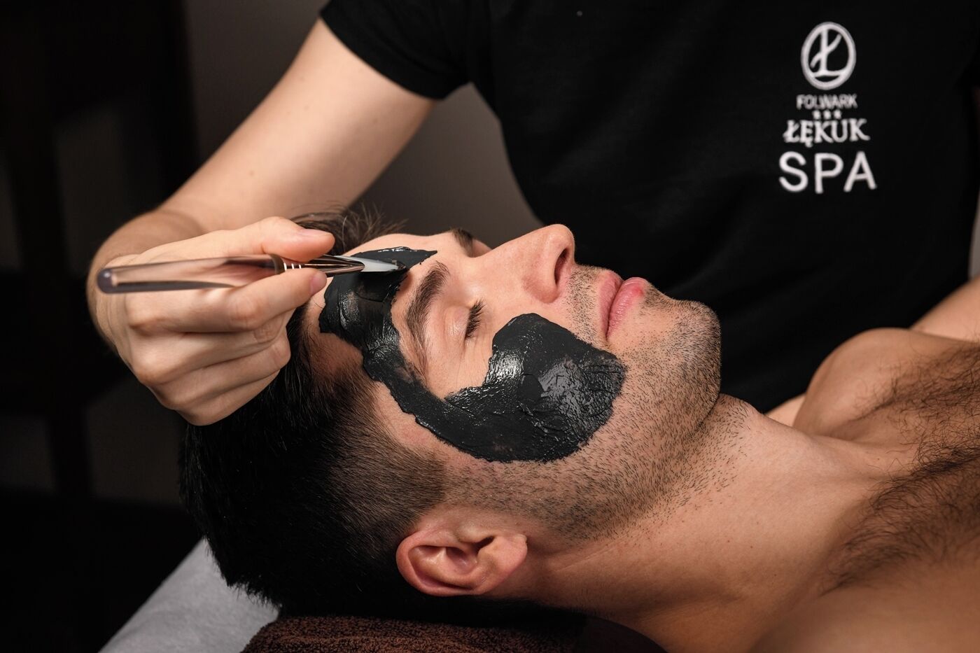A person receiving a facial treatment with a black mask, likely a spa service, indicated by the text on the attendant's shirt.