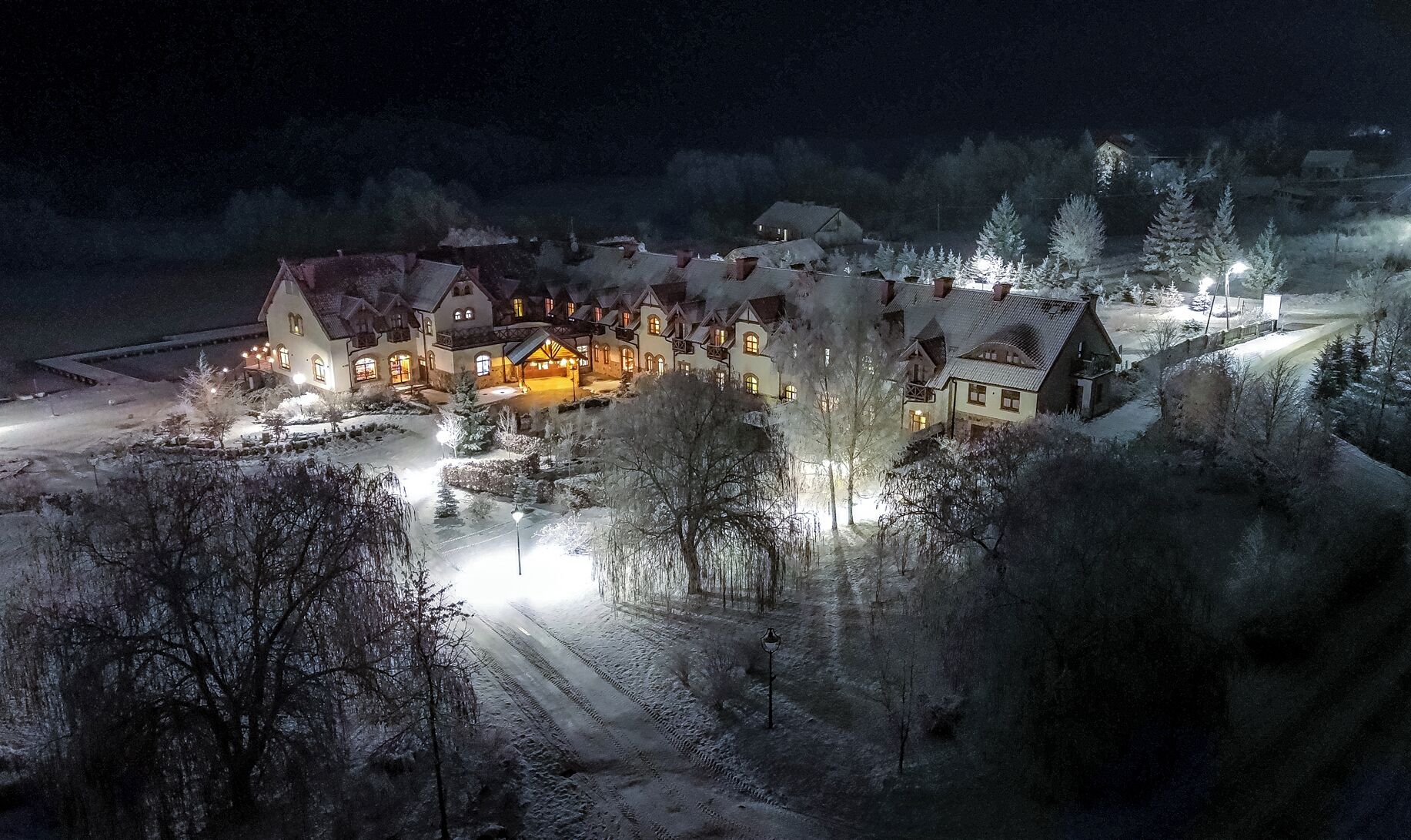 A snow-covered village with traditional houses, a main building with a lit entrance, and a clear road leading to it.