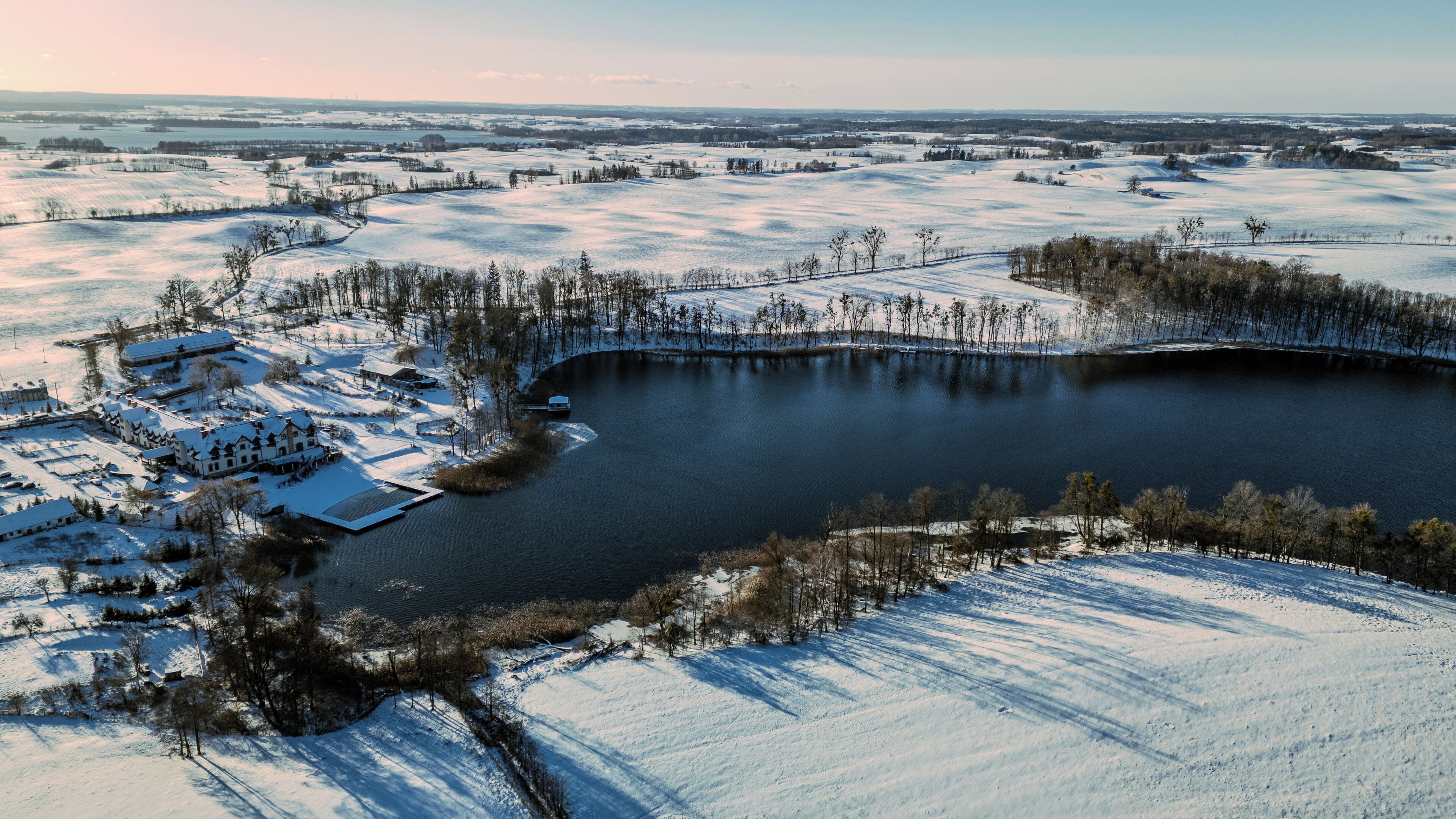 An aerial view of a lakeside property with snow-covered fields, a dock, and several buildings with snow on their roofs.