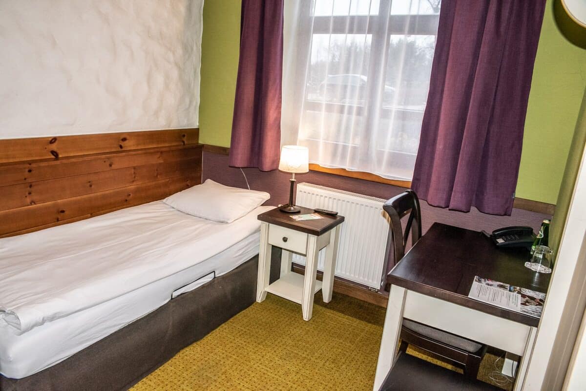 Una habitación de hotel pequeña con una cama individual, un escritorio con silla, una lámpara y una ventana con cortinas.
