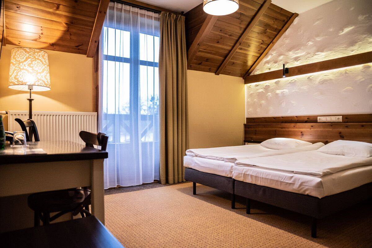 Una habitación de hotel con techo de madera, una cama con ropa blanca, un escritorio con lámpara y una ventana con cortinas.