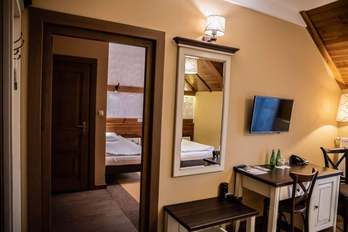 Habitación de hotel con puerta de madera, cama con ropa blanca, espejo, televisor, escritorio con silla y una pequeña mesa.