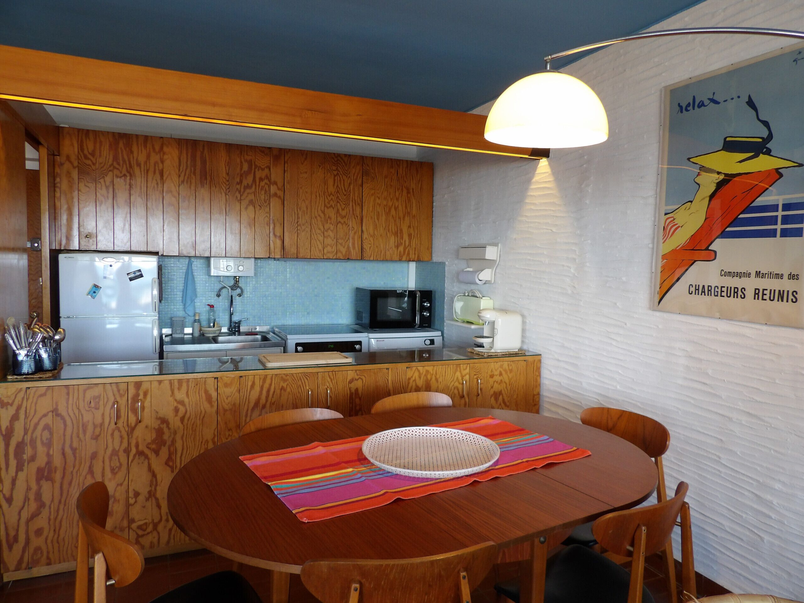 Cuina compacta amb armaris de fusta, nevera, microones i zona de dinar amb taula i cadires.