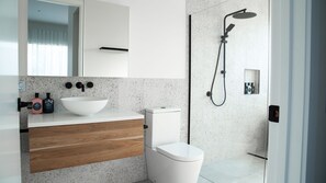 Un baño moderno con lavamanos blanco, tocador de madera, inodoro y ducha con cabezal de ducha manual.
