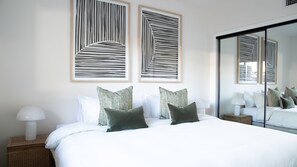 A modern bedroom with a large bed, two wall-mounted artworks, a mirror, and a bedside table with a lamp.