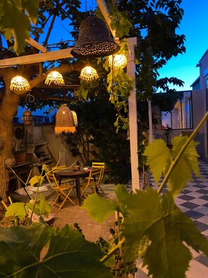 An outdoor patio area with a checkered floor, a wooden table with chairs, hanging lights, and a pergola with climbing plants. - Villa flower district 5 min walk from the beaches à Canet En Roussillon (Canet-en-Roussillon)