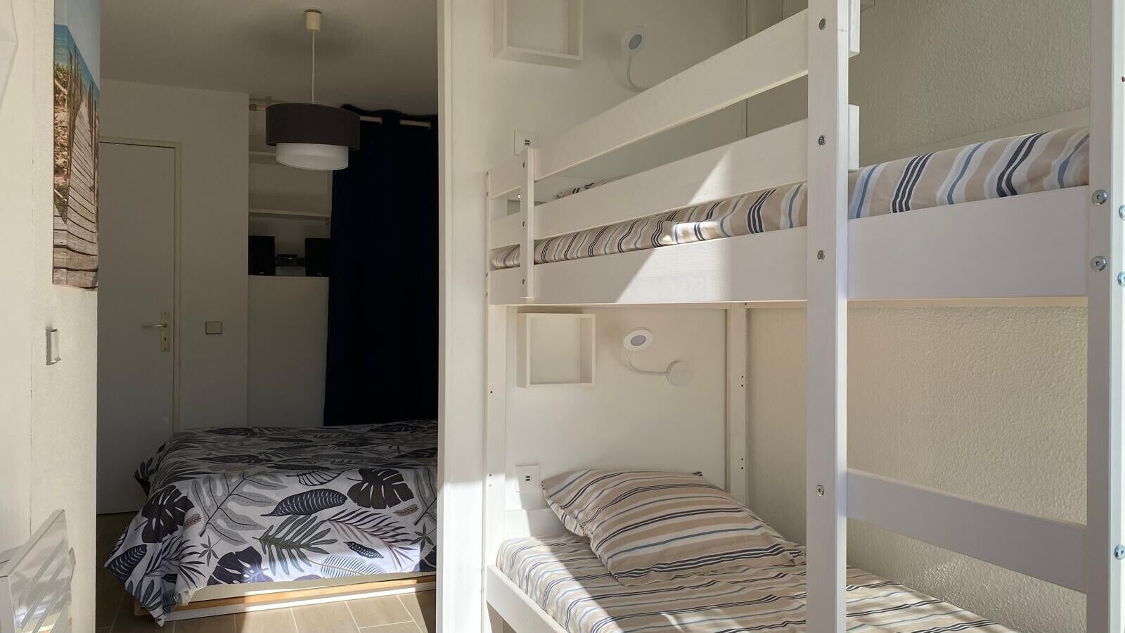 Een slaapkamer met een bed beneden en een stapelbed erboven. Er is een deur naar een andere kamer, een hanglamp en een wandlamp.
