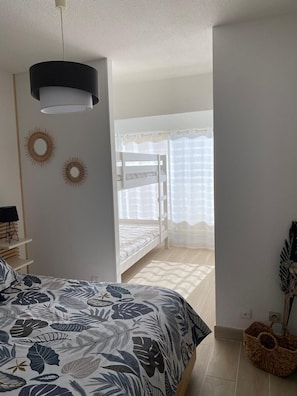 A bedroom with a bed, a hanging light fixture, a window with curtains, and a wall-mounted shelf.