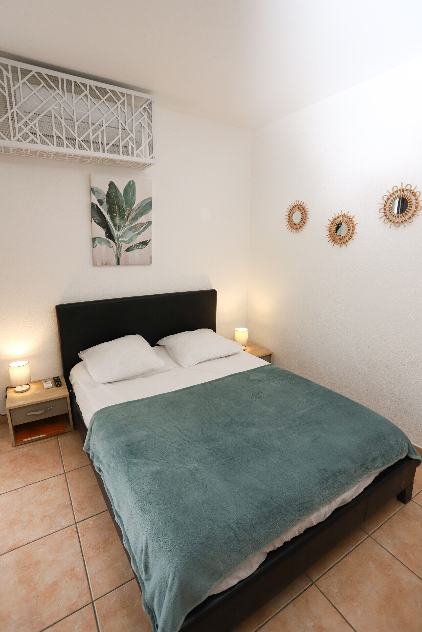 A single bed with a green blanket, a nightstand with a lamp, a wall-mounted plant, and two wall mirrors.