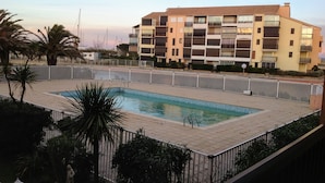 A rectangular swimming pool surrounded by a white fence, with a multi-story building in the background.