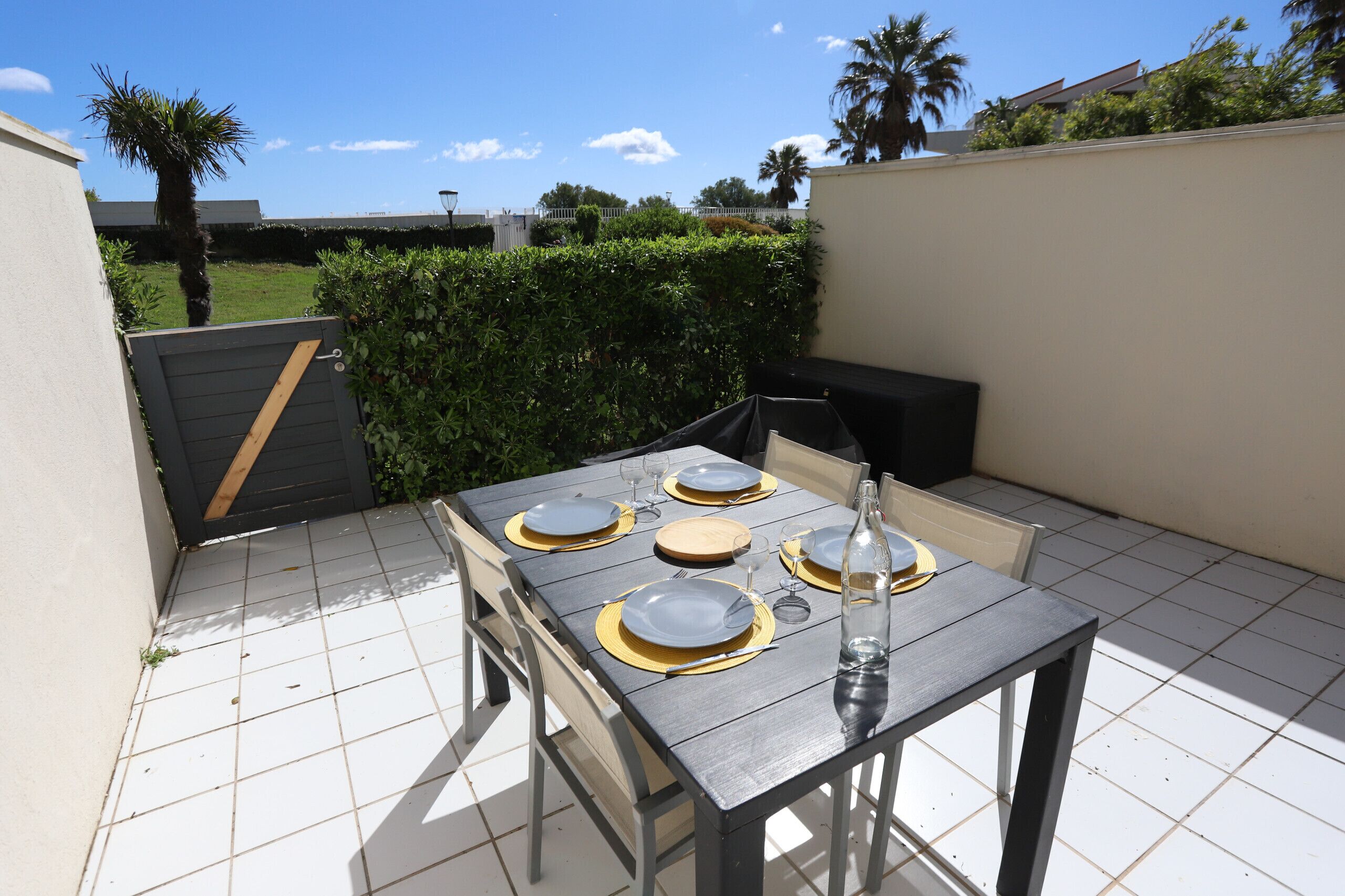 An outdoor patio with a table set for four, surrounded by chairs, and a large planter with greenery.