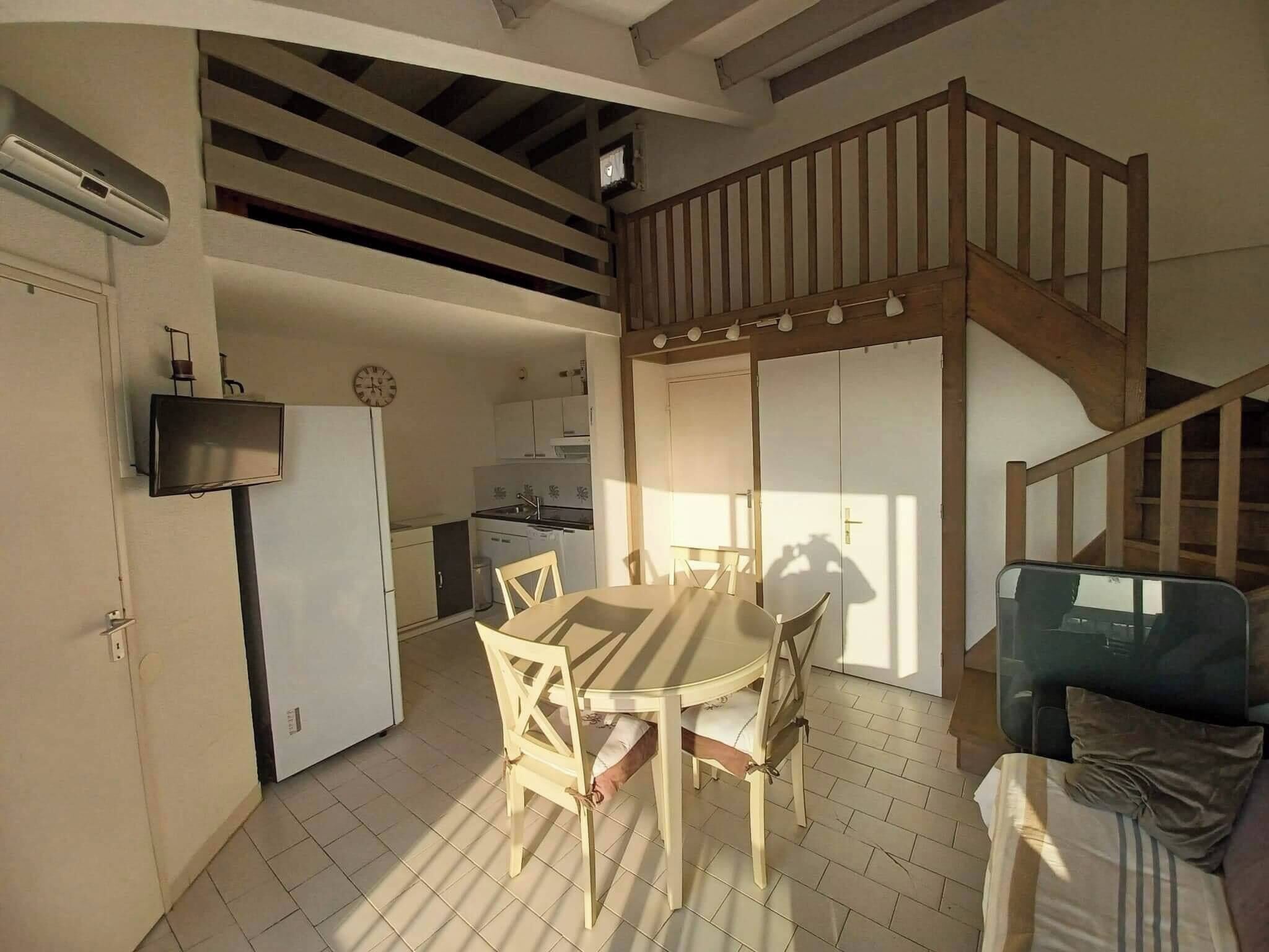 A spacious living area with a dining table, chairs, a sofa, a kitchen in the background, and a staircase.