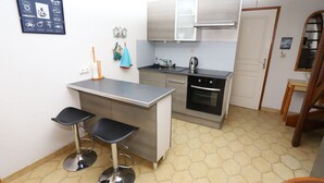 A compact kitchen with a central island, two bar stools, a built-in oven, and a small dining area with a wooden table and chairs.
