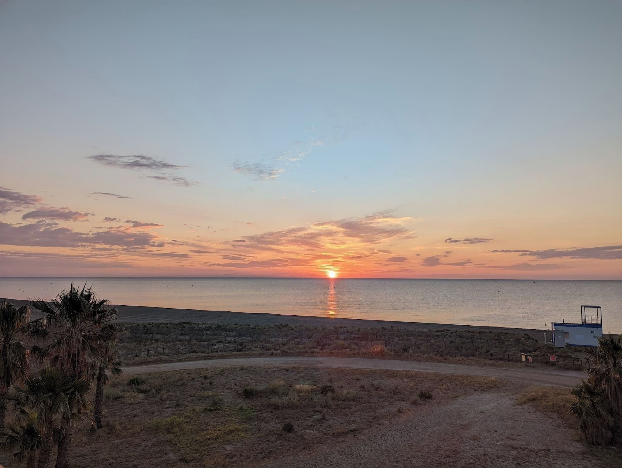 Un coucher de soleil sur une plage, avec un sentier de terre menant à une construction.