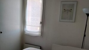 A small, neatly arranged bedroom with a single bed, a window with blinds, a framed picture on the wall, and a bedside lamp. - St Cyprien: Calm and gentleness on the edge of the lagoon (Saint-Cyprien)