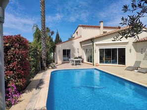 A swimming pool with a patio area, a covered seating area, and a building with a tiled roof.
