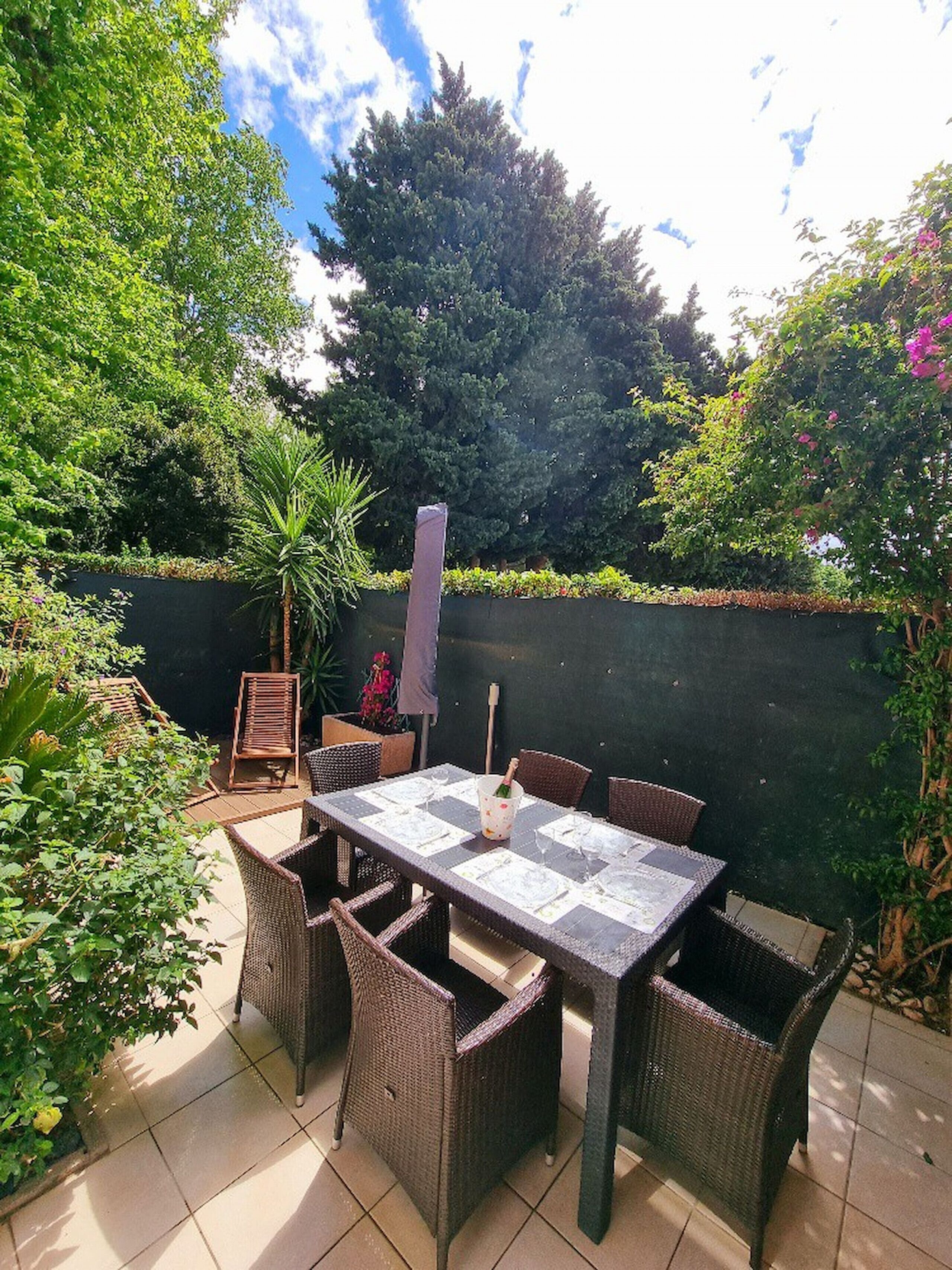 Un patio con una mesa dispuesta para cuatro personas, rodeado de sillas de mimbre, una planta en maceta y una planta alta al fondo.