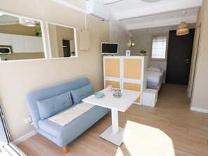 A compact living space with a blue sofa, a small white table, a TV, and a bed in the background.