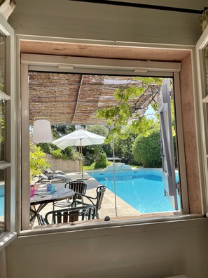A view from a window of a poolside area with a table and chairs, a covered seating area, and a swimming pool.