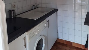 A compact kitchen with white cabinets, a black countertop, a built-in washing machine, and a clock on the wall. - St Cyprien beautiful holiday home near the beach (Saint-Cyprien)