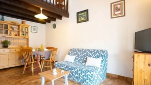 A cozy living room with a patterned sofa, wooden dining table, and chairs. A television is mounted on a wooden cabinet, and there are framed pictures on the wall. - St Cyprien beautiful holiday home near the beach (Saint-Cyprien)