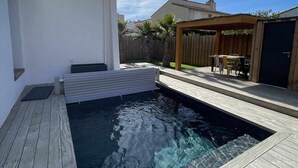 A swimming pool with a wooden deck, a covered seating area, and a white building in the background.