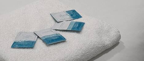 A neatly made bed with a white pillow and a white towel. On the towel, there are three Carell shower gel packets.