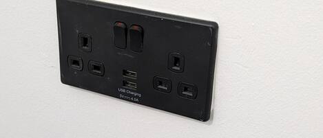 Un enchufe eléctrico negro con puertos de carga USB y de 5V en una pared blanca.