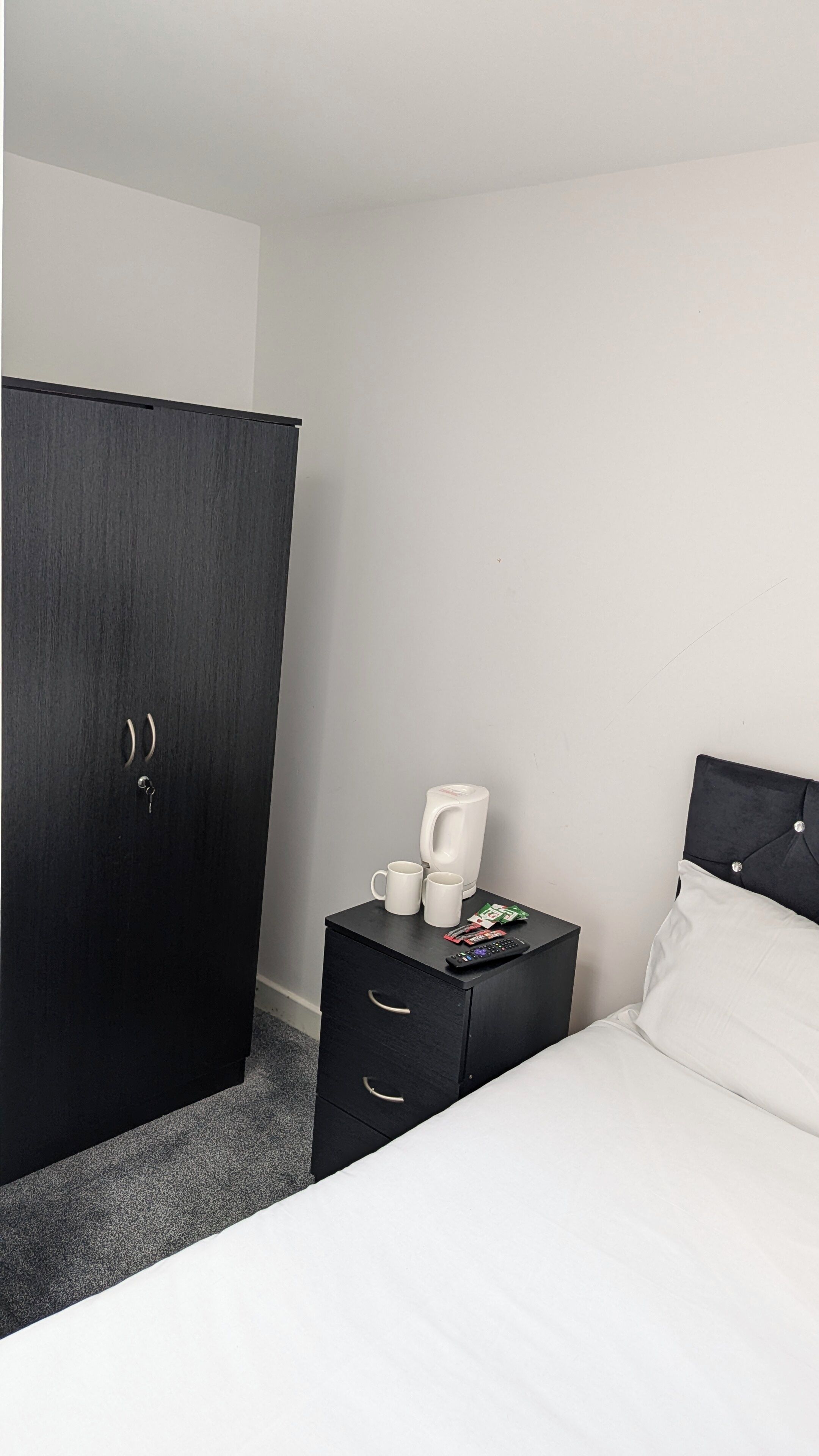 A hotel room with a bed, a nightstand with a coffee maker, and a wardrobe.