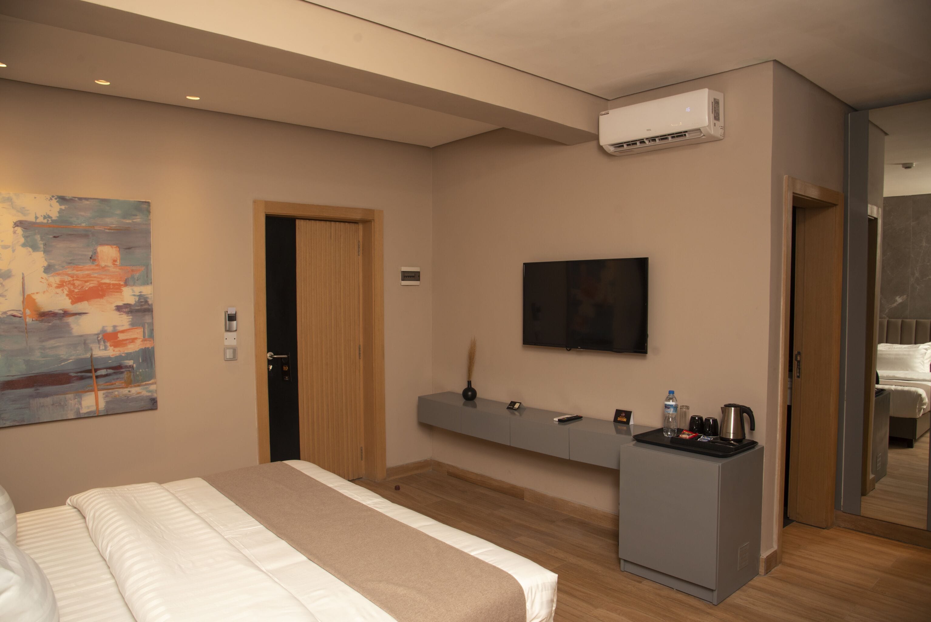 A modern hotel room with a bed, a mounted television, a small desk with a kettle, and a painting on the wall.