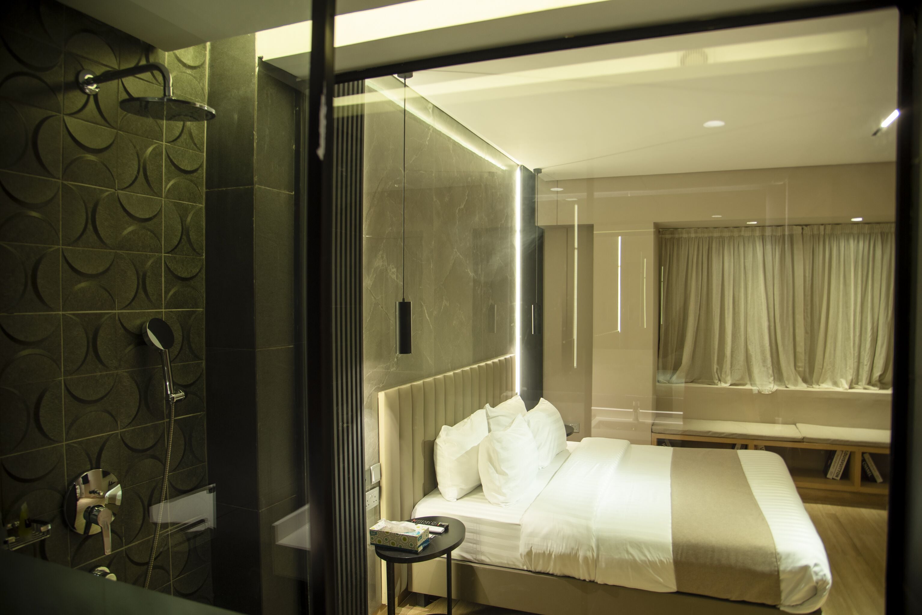 A modern hotel room with a large bed, a bedside table, a shower area, and a view of the city through a window.