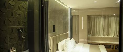 Habitación de hotel moderna con una cama grande, una mesita de noche, zona de ducha y vistas a la ciudad a través de una ventana.