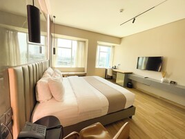 Habitación de hotel con cama, mesita de noche, silla, escritorio, televisión y ventana con cortinas.