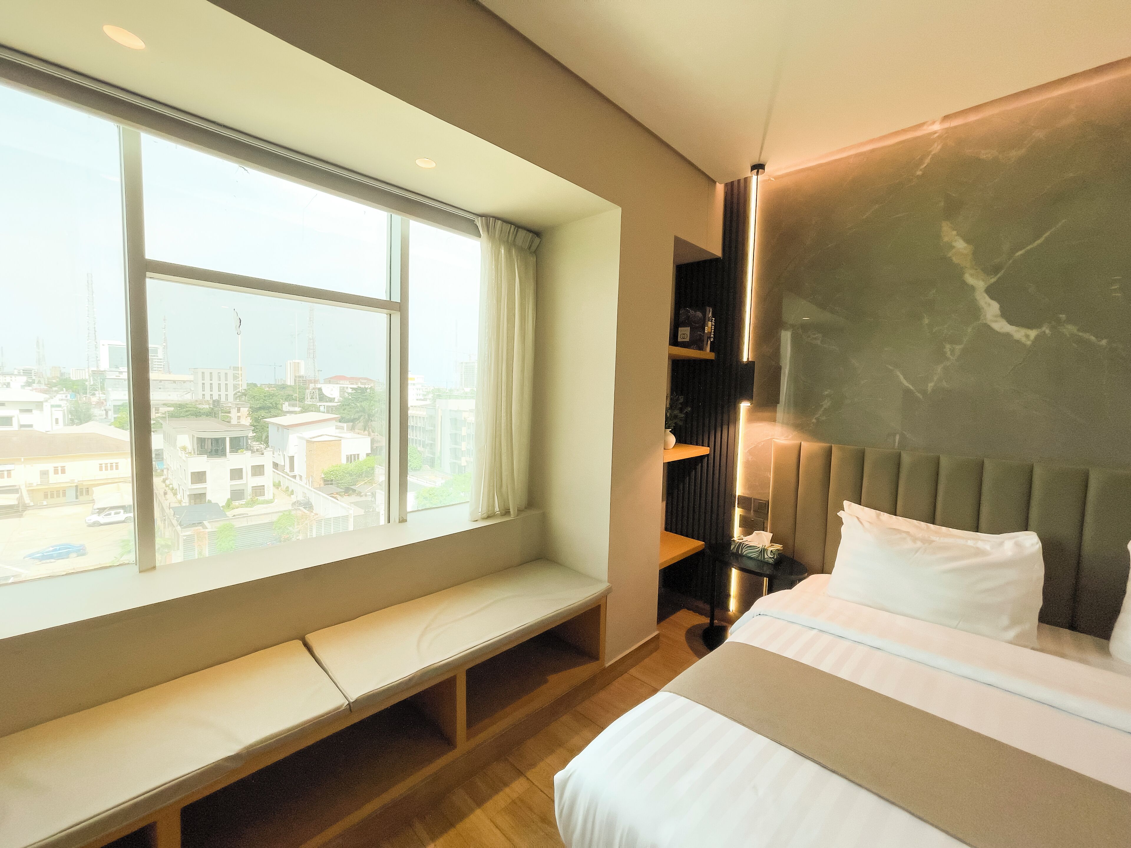 Une chambre d’hôtel moderne dotée d’une grande fenêtre, d’un banc, d’un lit avec une tête de lit et d’une étagère avec des livres et une plante.