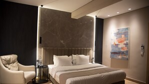Un dormitorio moderno con una cama grande, cabecero, mesita de noche, una silla y una obra de arte colgada en la pared.